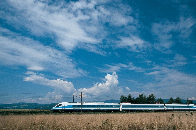 Image of a high-speed Slovenian train darting through grassland under a cloudy blue sky